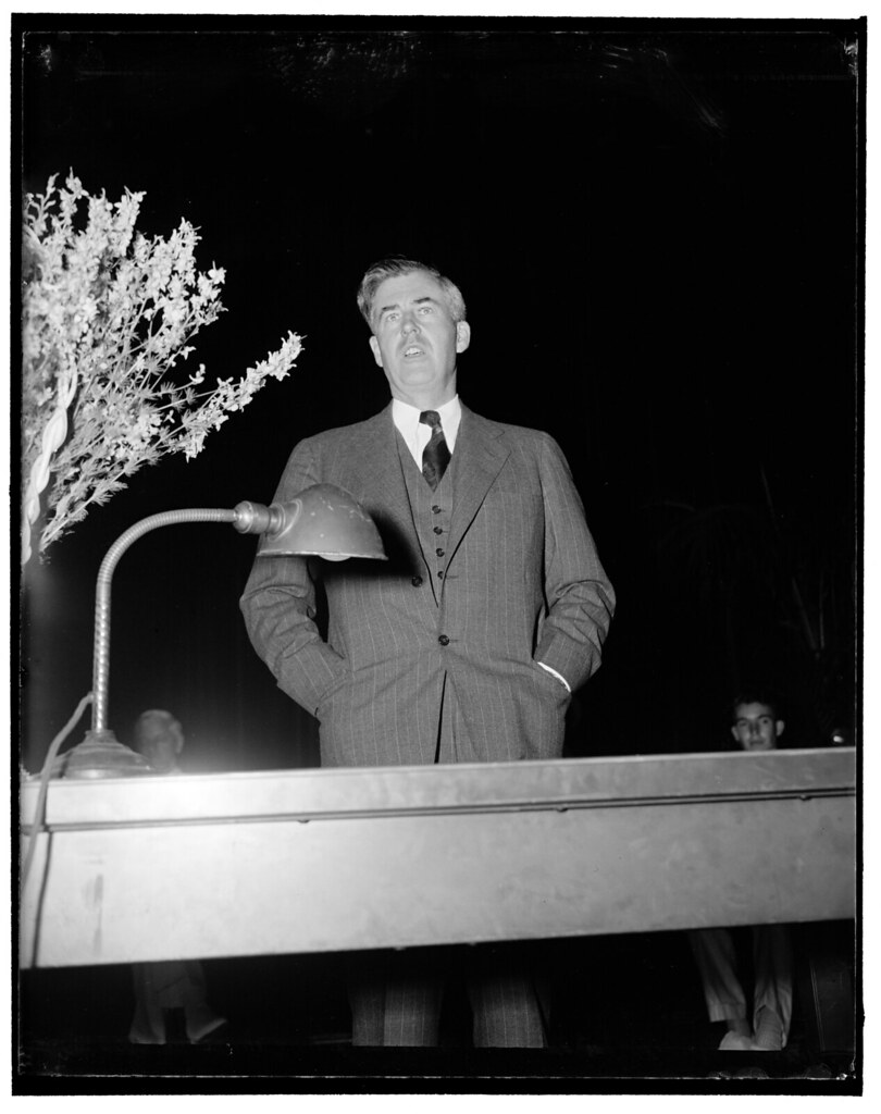 A black and white photograph of Henry Wallace speaking with his hands in his pockets